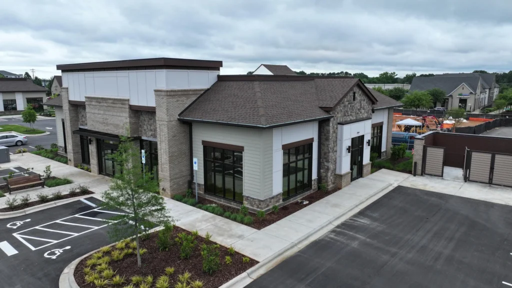 Aerial view of a modern retail building with brick and stone facade, large glass storefronts, and a parking lot with handicap spaces nearby.
